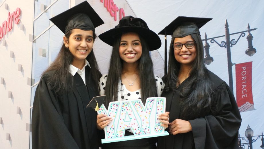 Three graduates wearing their caps and gowns standing in front of a photo of the Buhler centre.