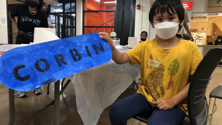 A boy with a art project sign with his name Corbin in blue.
