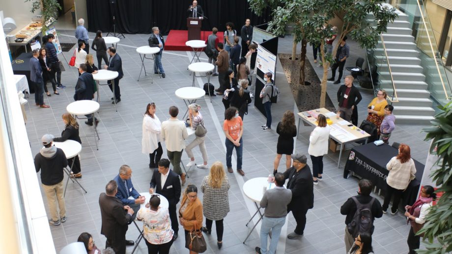 Photo of 45 people networking in the atrium of the Richardson College for the Environment and Science Complex. Centered at the top of the photo, Vice-President, Research and Innovation, Jino Distasio addresses the crowd. In the top right corner of the photo are science research posters with student presenters.