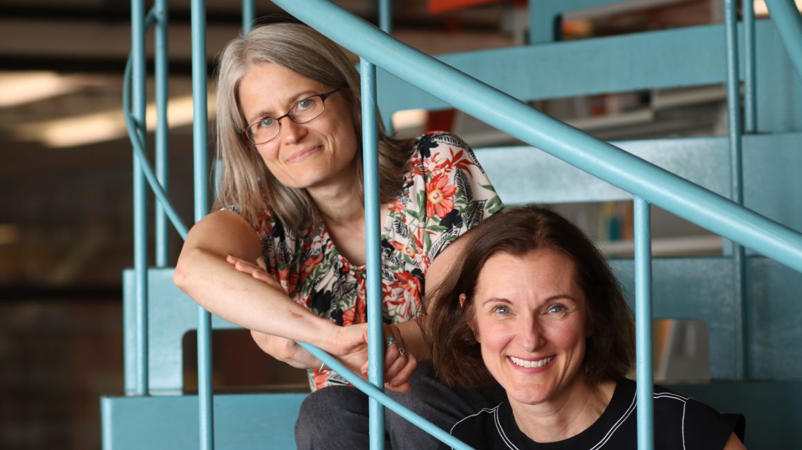 Dr. Tracy Whalen and Dr. Anne-Laurence Caudano sitting on the spiral staircase in the UWinnipeg Library.