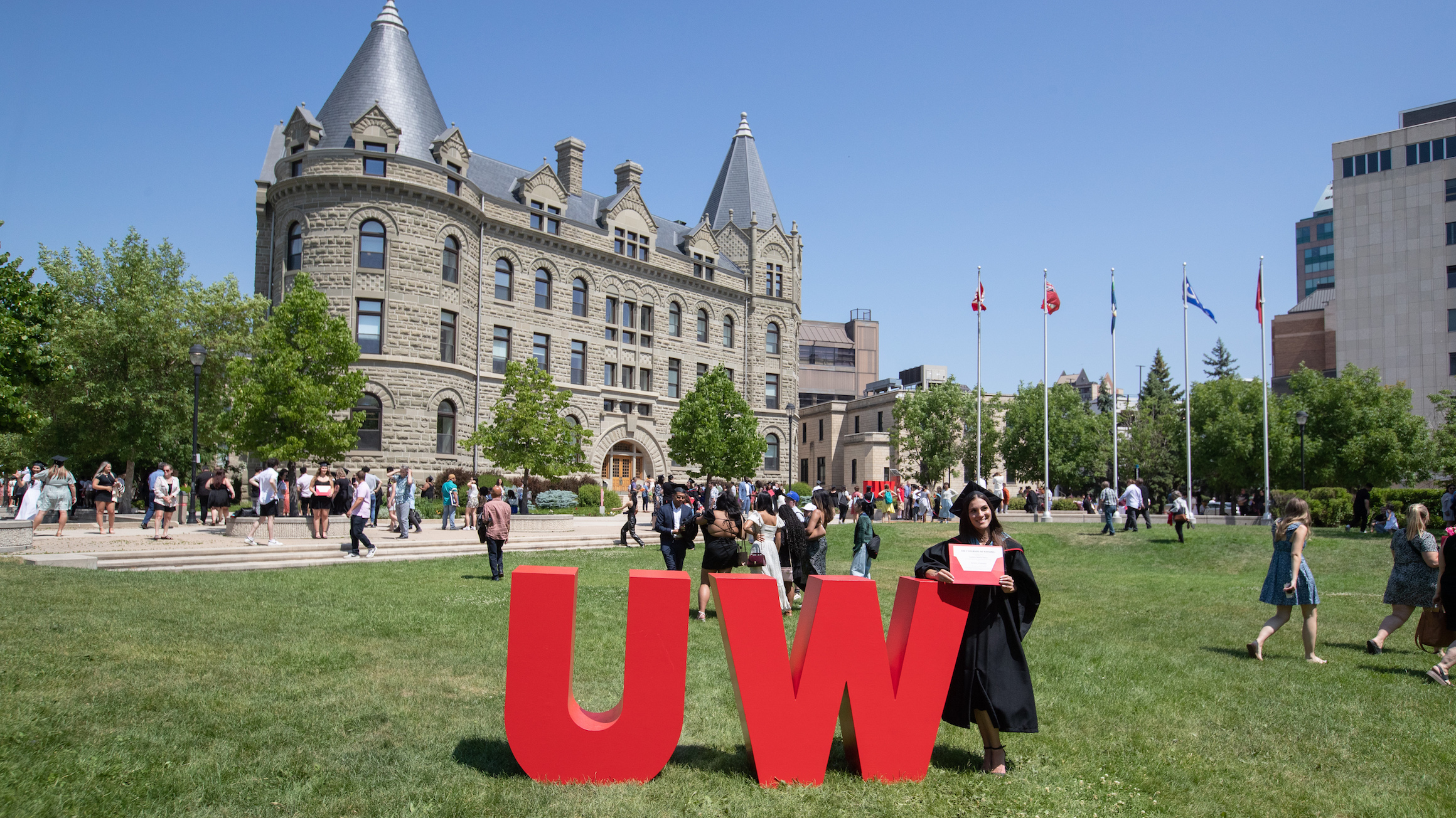 A graduate poses for a photo next to the UW sign on the front lawn of campus. Wesley Hall is in the background.