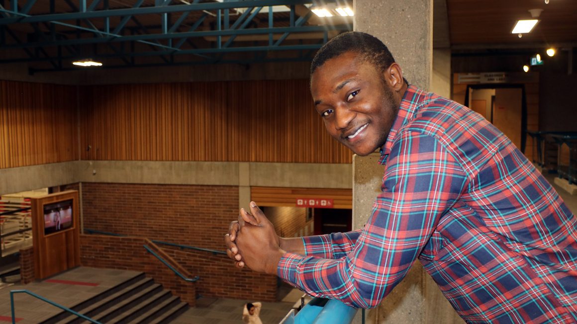 Arnold Osei in red and blue plaid shirt smiles at the camera while leaning on a railing above a large room with wood and brick walls.