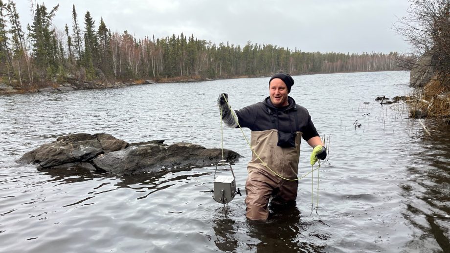Dylan McKenzie stands waist-deep in Hamell Lake collecting sediment samples during an October 2023 research trip to Flin Flon.