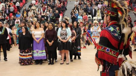 Gathering of a group of people wearing a mix of contemporary and traditional Indigenous garments stand in front of a crowd.