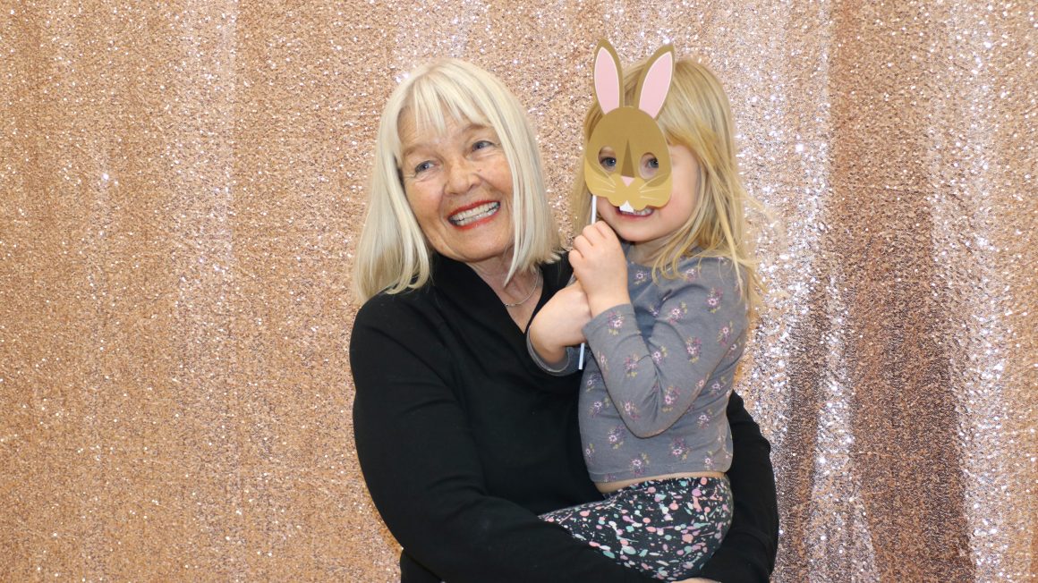Child holds a bunny mask in front of her face and is held up by her grandmother in front of a sparkling backdrop.