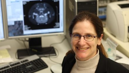 Headshot or Dr. Melanie Martin in front of computer with MRI image on screen 