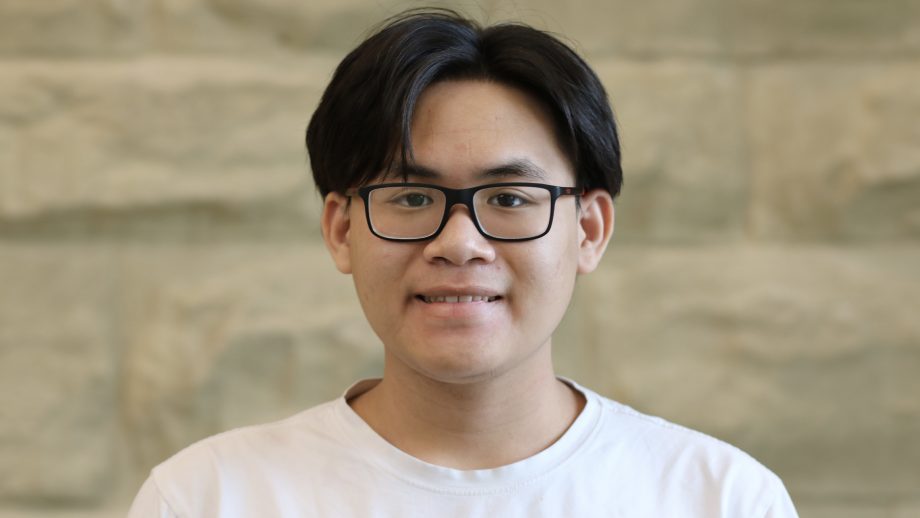 UWinnipeg's international student Collegiate student,from Vietnam Pham Tran Phu, wears a white t-shirt and stands against a stone backdrop in Leatherdale Hall.