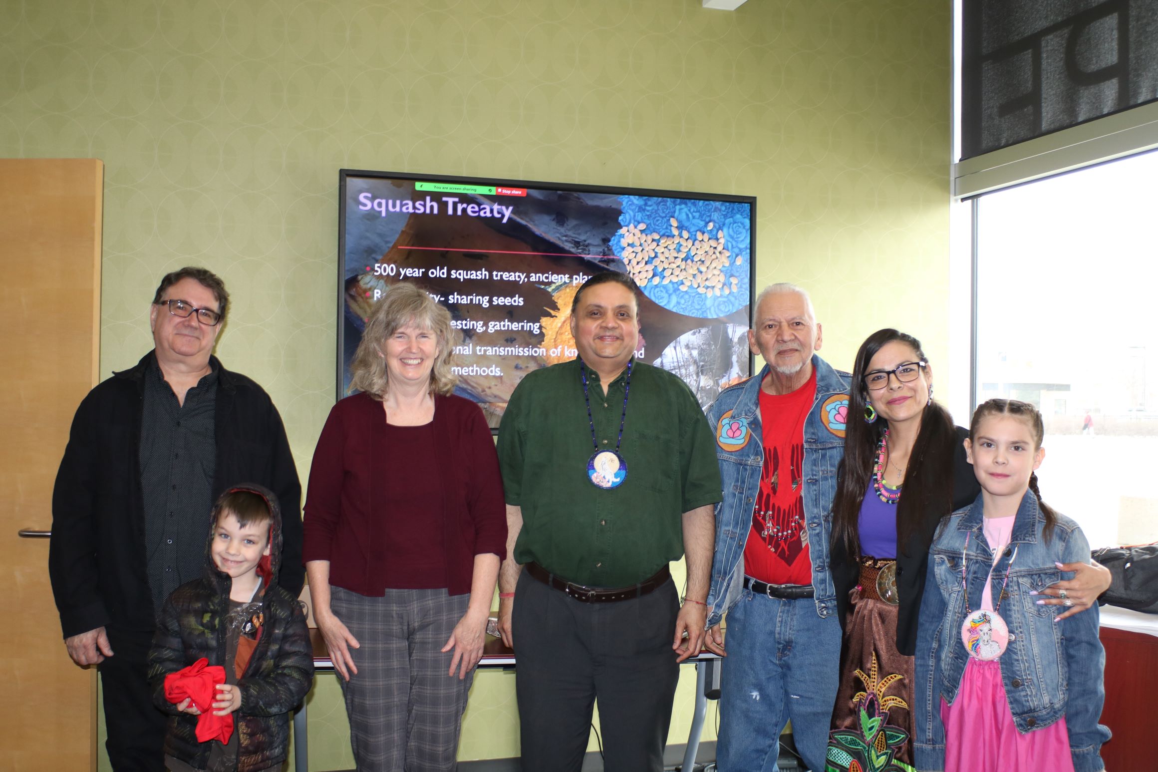 Dr. Mark Ruml, Dr. Shirley Thompson, Dr. Shailesh Shukla, Knowldege Keeper Dave Daniels, Anna Neil, and two children stand in a classroom with a video screen behind them.