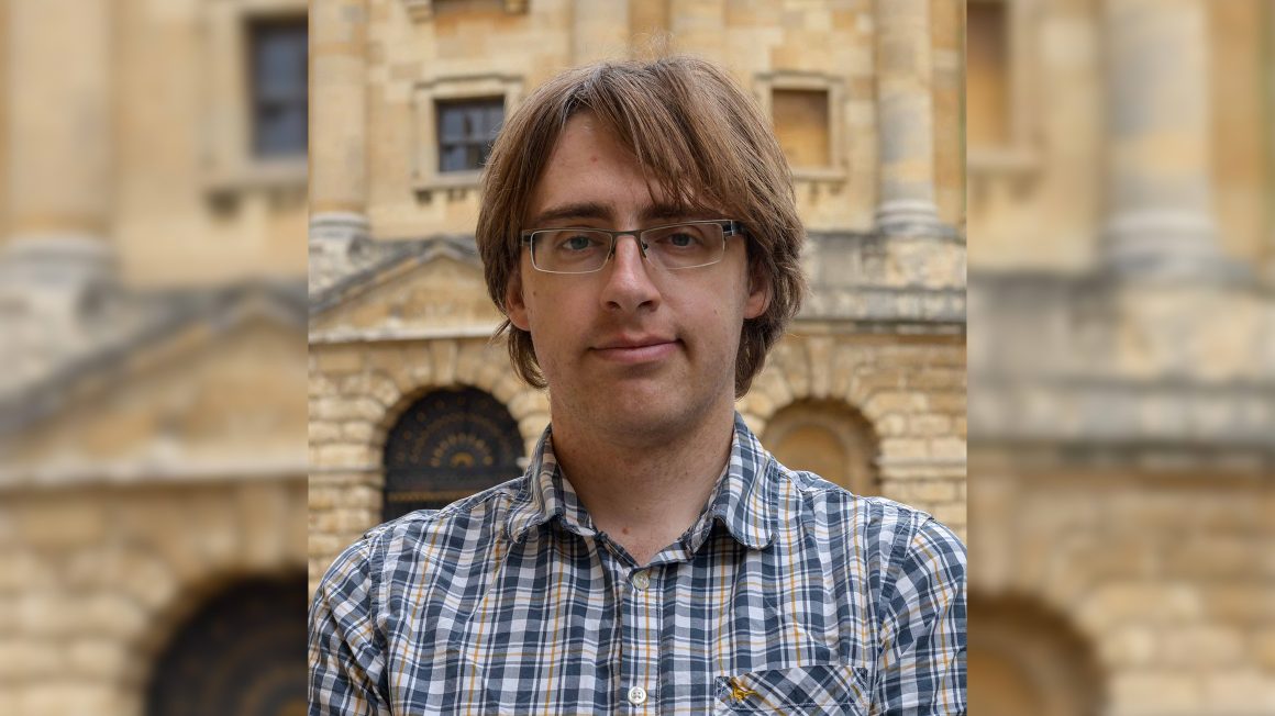 A man in a plaid shirt stands in front of a stone building.