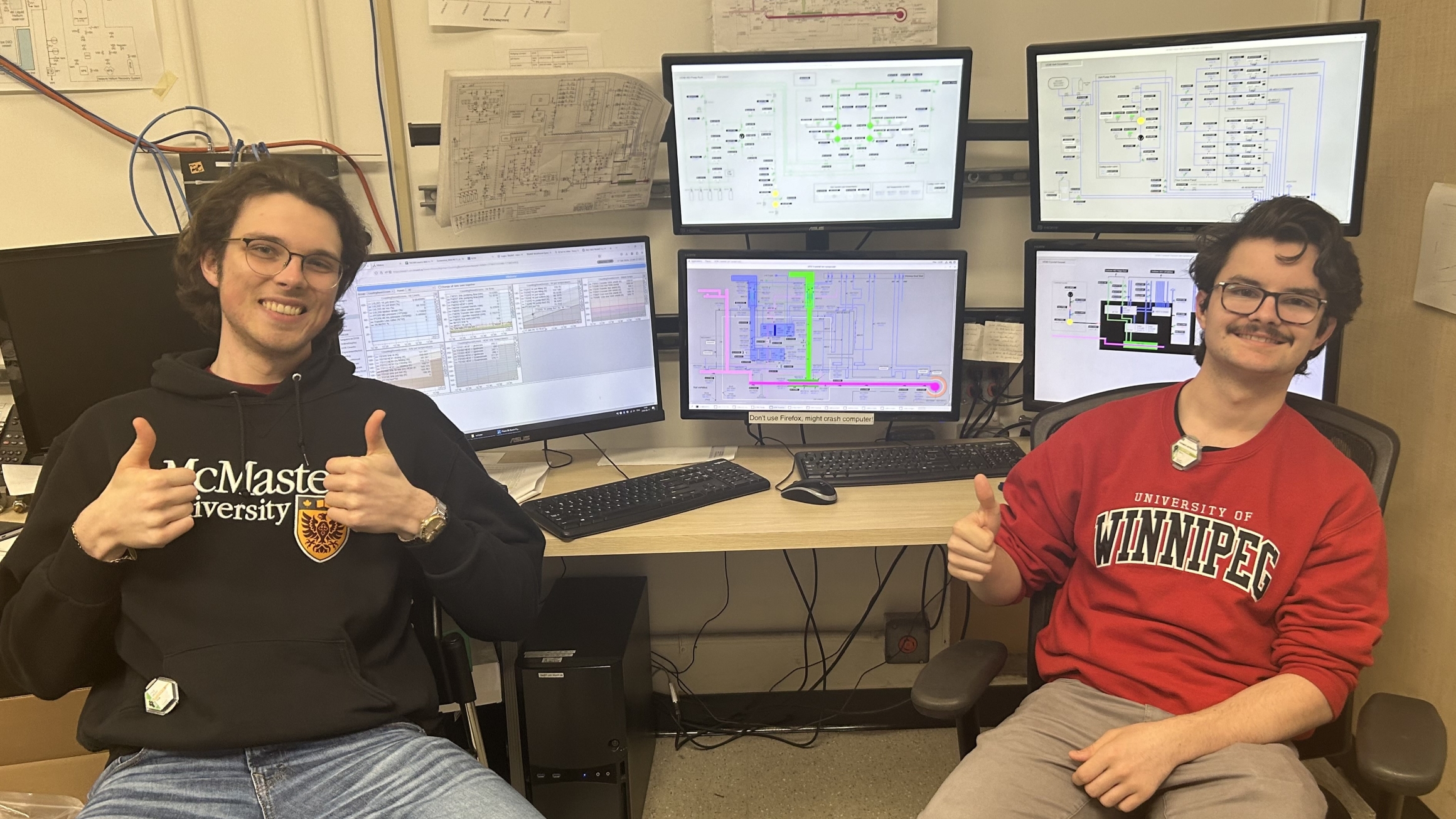 Two students, seated and wearing university sweaters, make a thumbs-up gesture in front of a bank of computer monitors.