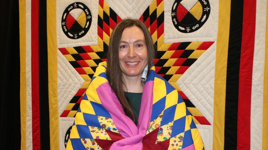 Kate Binesigichidaakwe McDonald wrapped in a blanket standing in front of a star blanket.