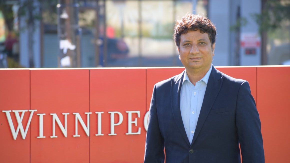 Dr. Anuraag Shrivastav stands next to a red University of Winnipeg sign