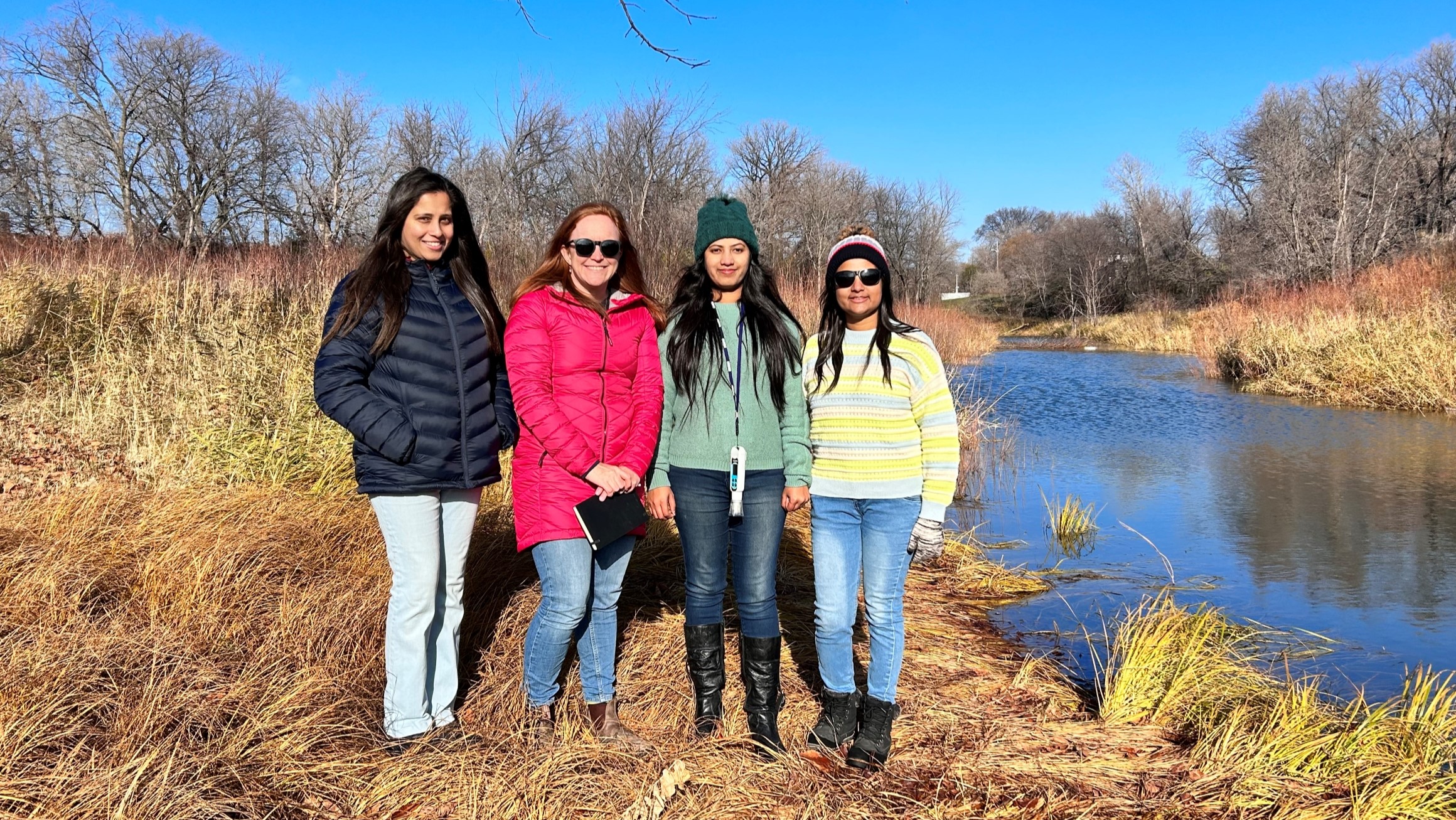 4 people stand in front of a creek in autumn.