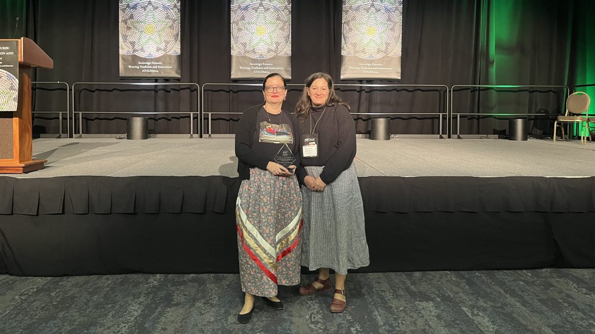 Two women standing in front of a stage hold an award.