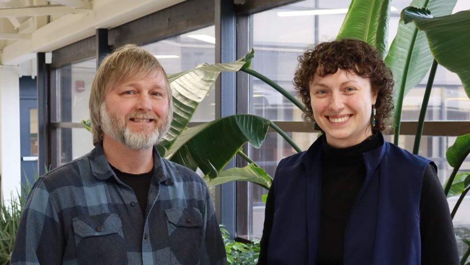 In a hallway with plants and windows, a man with light brown hair and a beard stands next to a woman with dark brown hair.