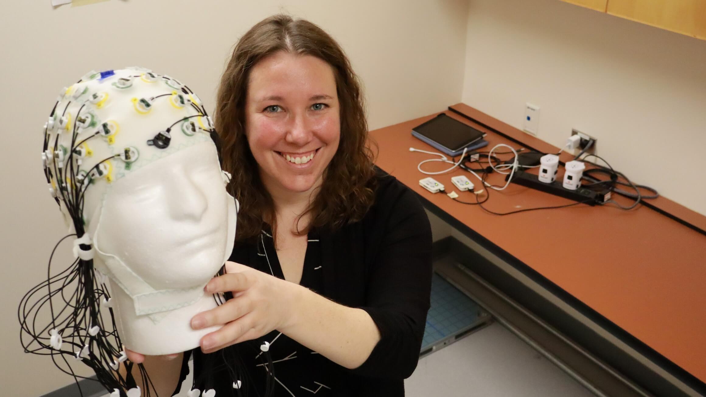 A woman with brown hair holds a styrofoam skull wearing a cap covered in electrodes and wires.