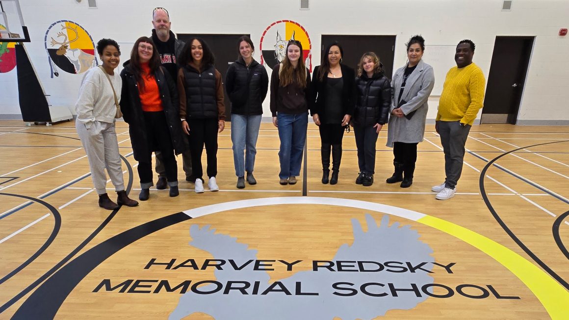 Mozit Tsegai, Haley Carr, Wayne Davies, Kyanna Wonnacott, Christina Duerksen, Elise Stokes, Sheila North, Chloe Heidinger, Cynthia Jourdain (Bimose Tribal Council), Claude Pike (Principal of Harvey Redsky Memorial School) standing in a gym.