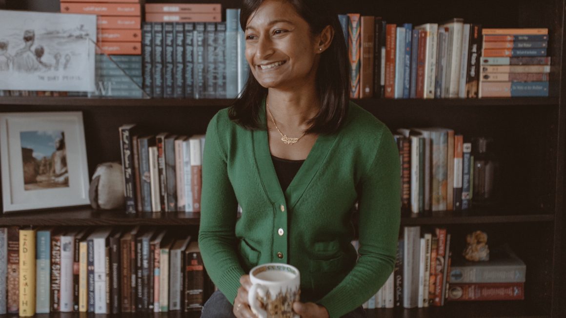 writer Sharon Bala standing in front of shelves of books, holding a mug.