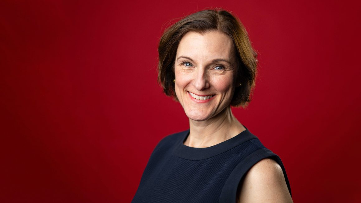 A woman with brown hair stands in front of a red backdrop.