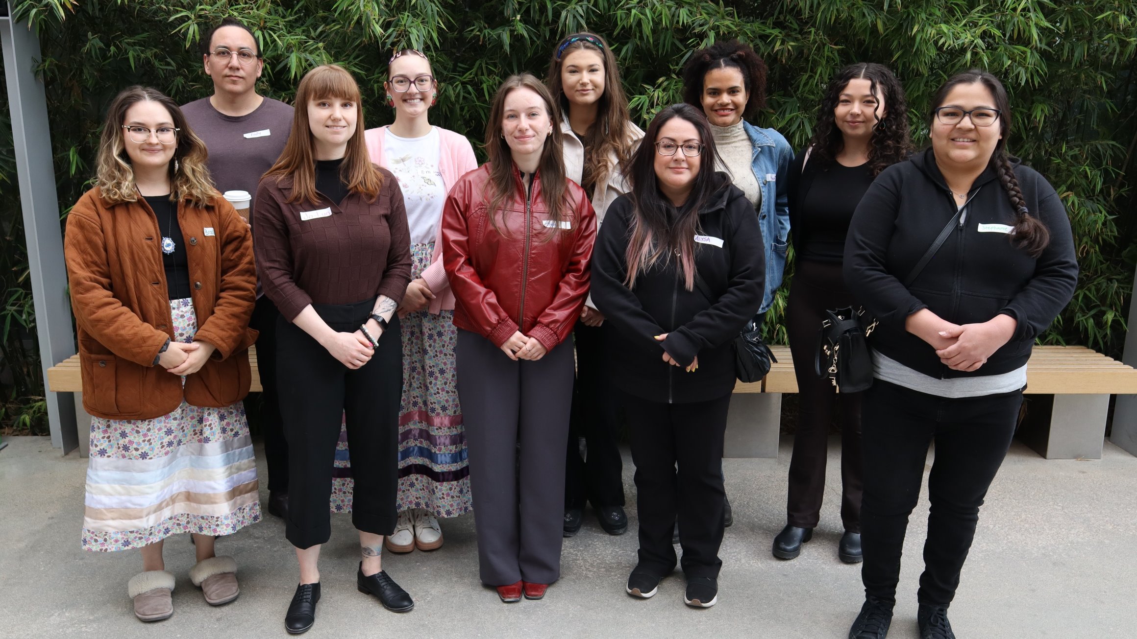 A group of 10 students stands in a row in front of a wall of greenery.