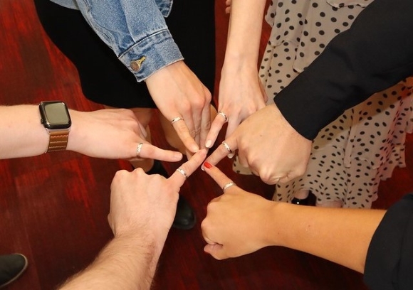 Six hands in a circle displaying silver rings on pinky fingers.