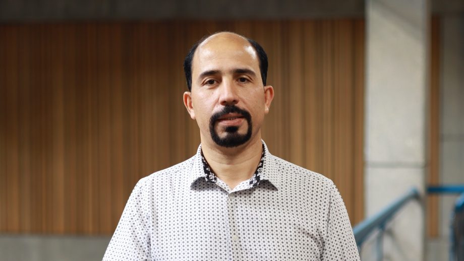A male university professor in a button-up shirt stands in front of a wall with wood panelling.
