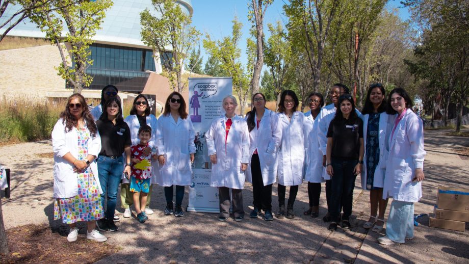 A group of scientists in white lab coats gathers for an outdoor photo under some trees.