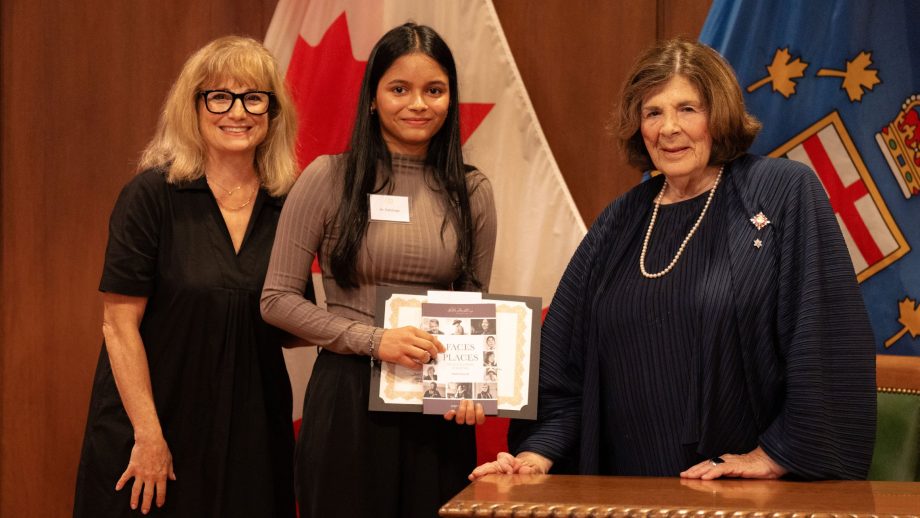 Two women present a student with a scholarship in front of a row of flags.