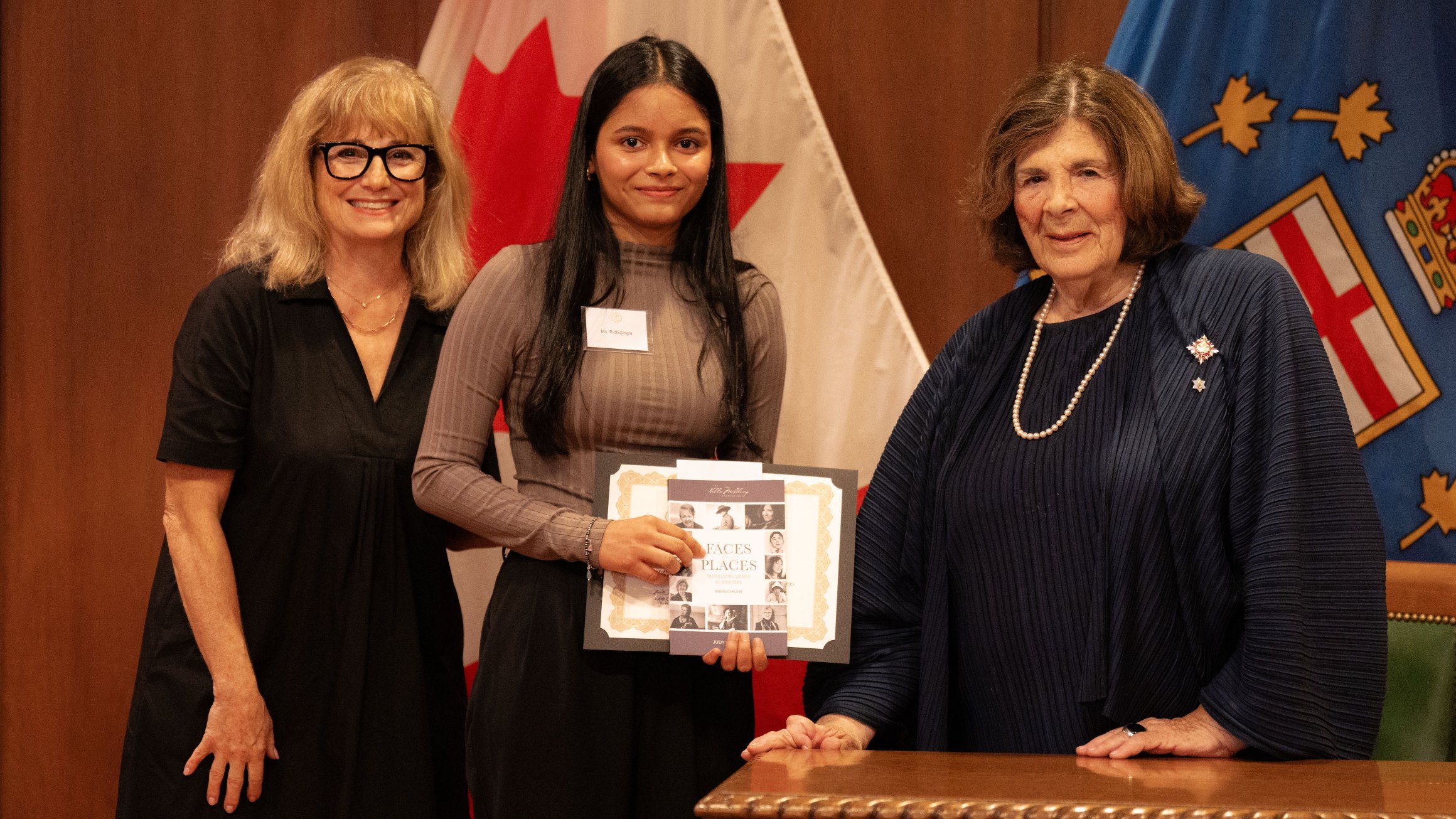 Two women present a student with a scholarship in front of a row of flags.