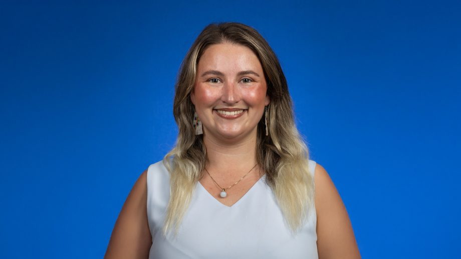 A student with long hair and wearing a white top stands in front of a blue backdrop.