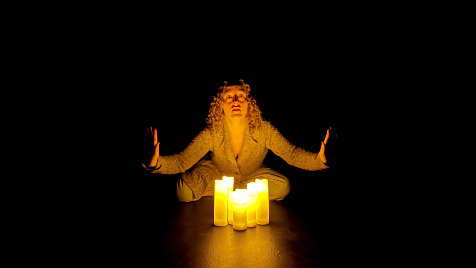 Actor Elli Suppes sits on a dark stage with lit candles.