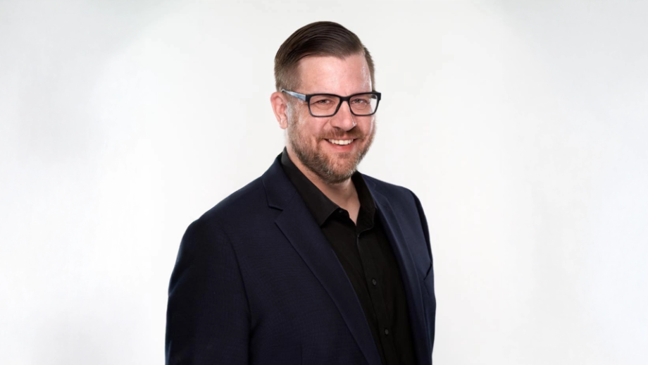 A man with brown hair, glasses, and a beard smiles in front of a white backdrop.