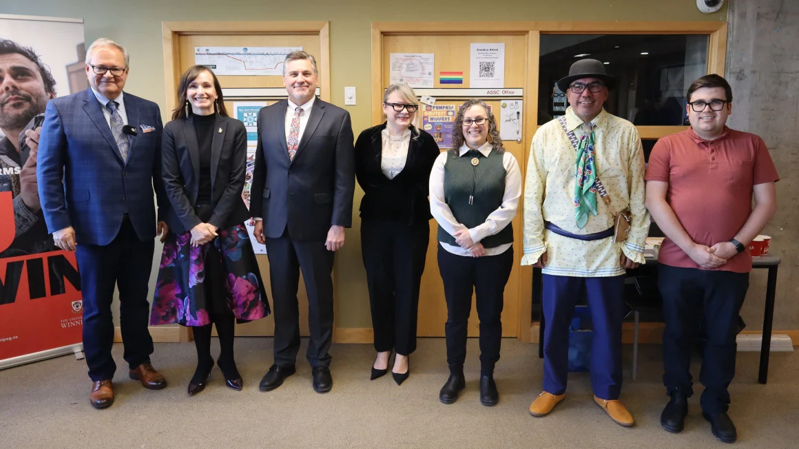 Seven individuals stand in a row at the front of a room.