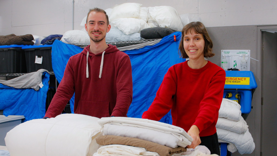 A man in a red hoodie and a woman in a red sweater fold smile as they fold laundry on a table in a commercial laundry facility.