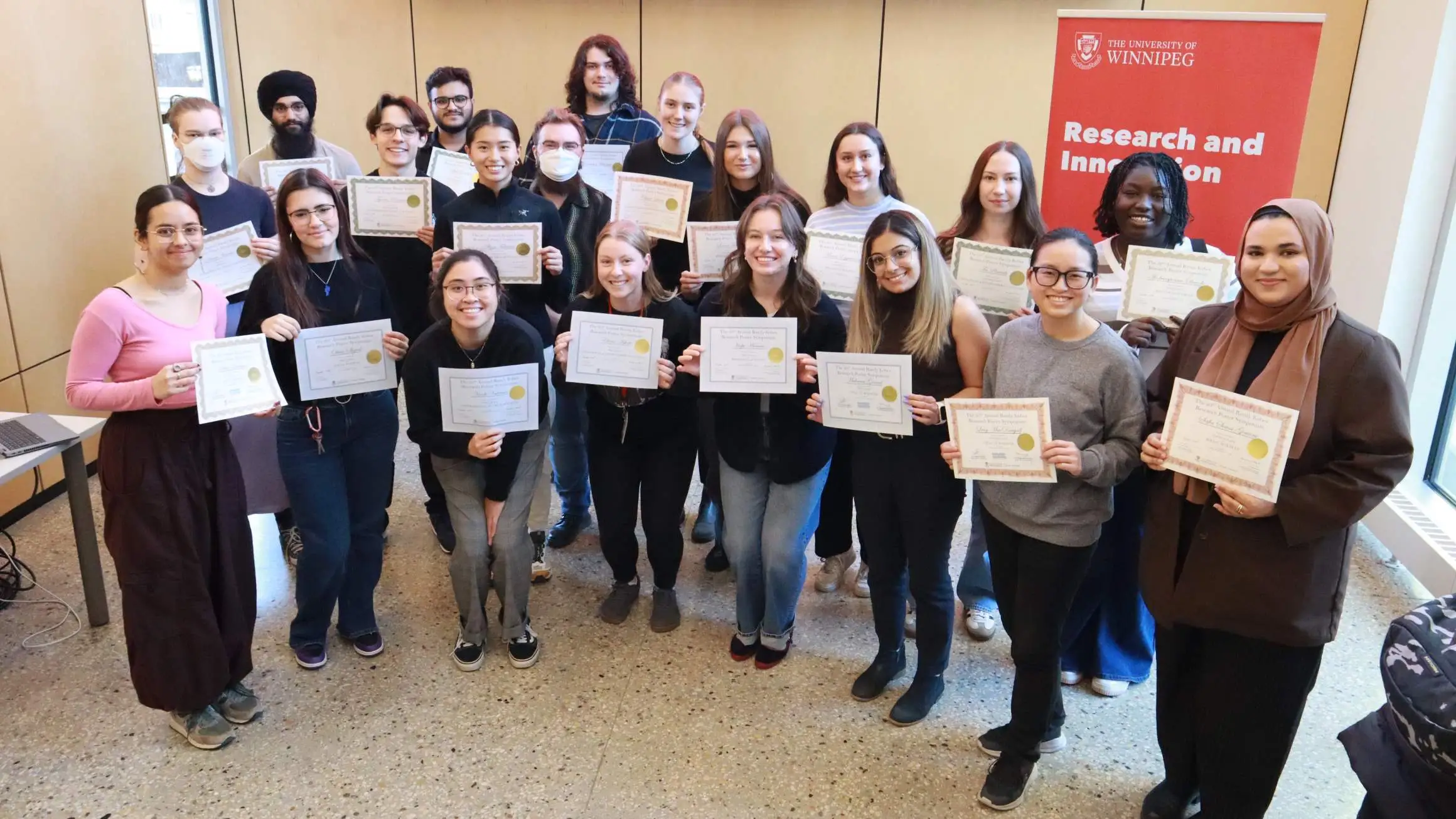 Twenty students stand in a group at the front of a room displaying certificates.