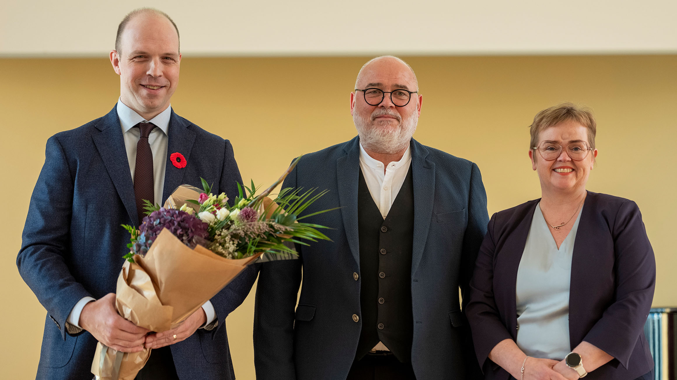 Dr. Ryan Eyford holding a bouquet of flowers standing next to Logi Einarsson, Iceland’s Minister of Culture, Innovation, and Higher Education and Silja Bára R. Ómarsdóttir, Rector (i.e., President) of the University of Iceland.