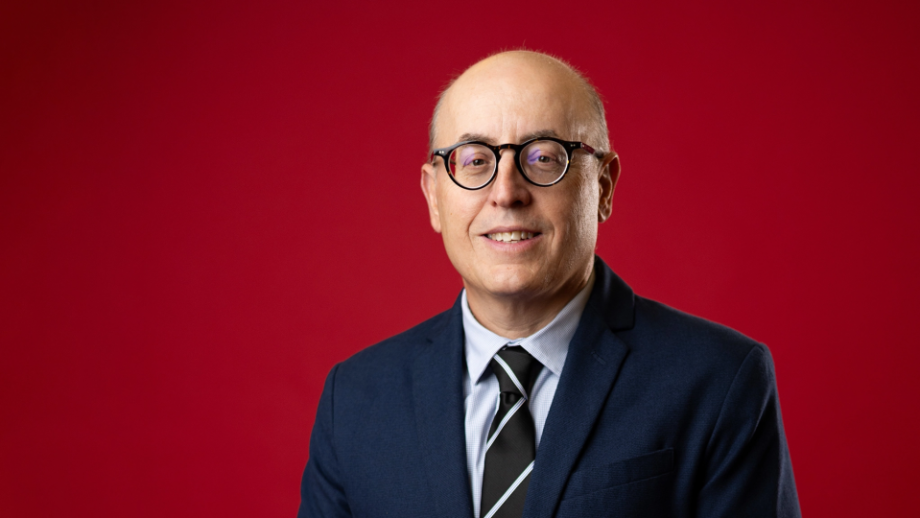 A man with glasses in a dark suit and striped tie is pictured against a red backdrop.