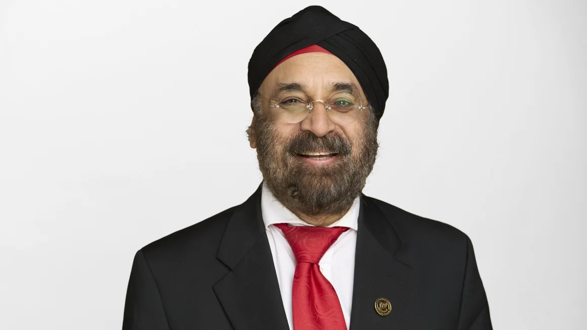 A man with glasses, a beard, and turban wears a suit and smiles in front of a white studio backdrop.