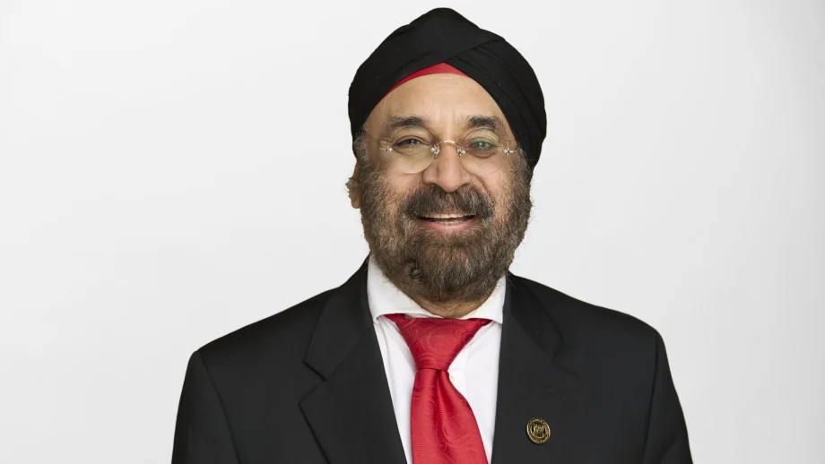 A man with glasses, a beard, and turban wears a suit and smiles in front of a white studio backdrop.