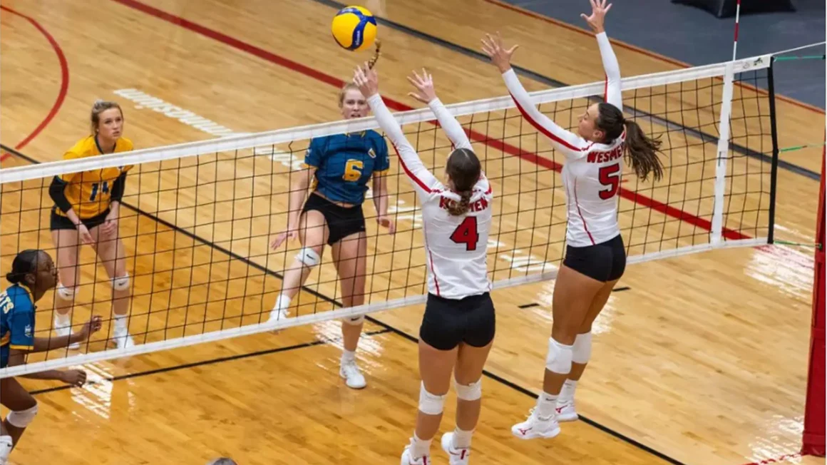 Two women's volleyball players in white jerseys jump for a ball at the net.