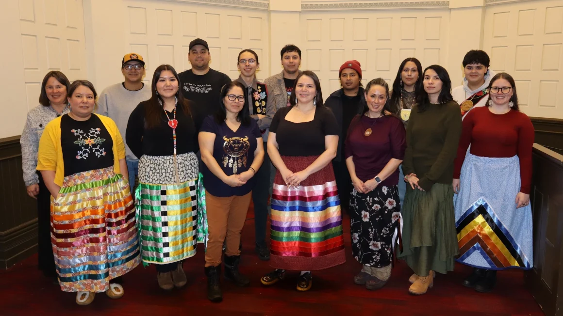 A group of Indigenous students and faculty gather for a group photo in a room with white walls and red carpet.