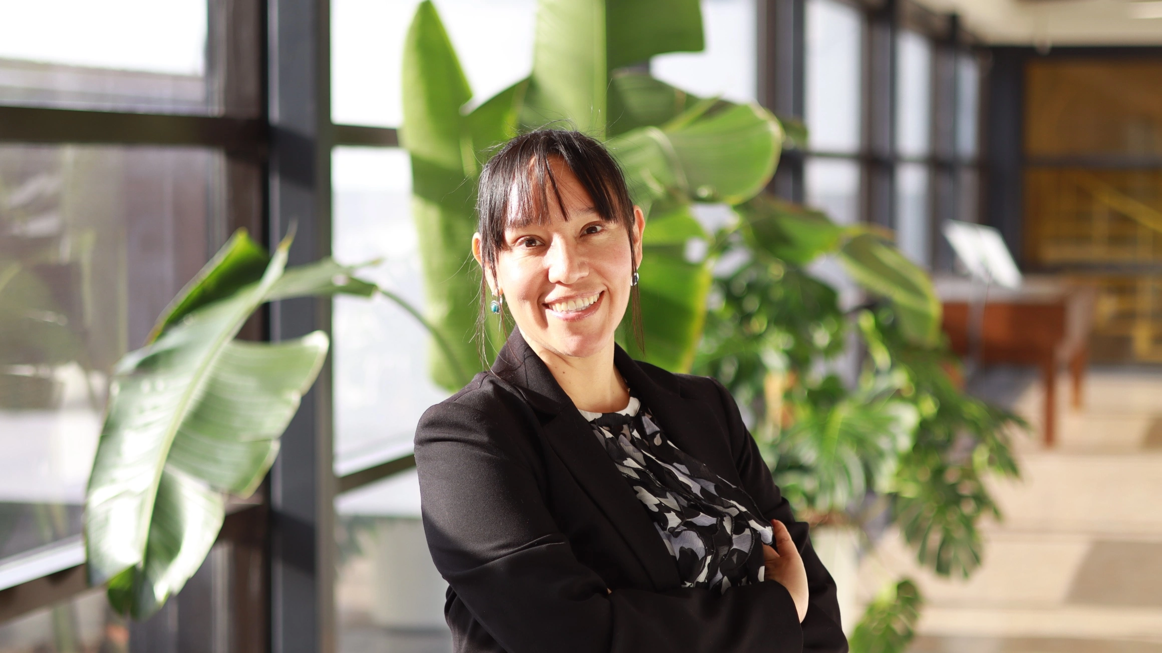 A woman with brown hair and a black top stands in a sunlit hallway with plants.
