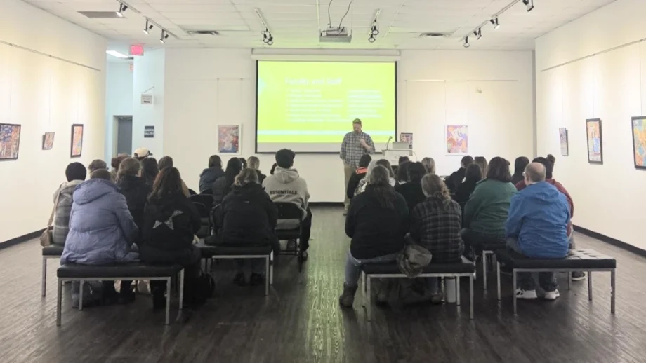 A man stands at the front of a large gallery-style room and presents in front of a drop-down screen. Seated facing him are about 40 people listening to the presentation.