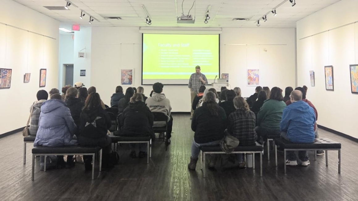A man stands at the front of a large gallery-style room and presents in front of a drop-down screen. Seated facing him are about 40 people listening to the presentation.