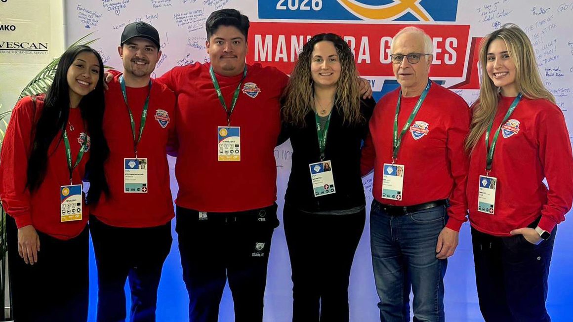 Six people, a mix of students and faculty members, stand in a row wearing red tops and lanyards at the Manitoba Games.
