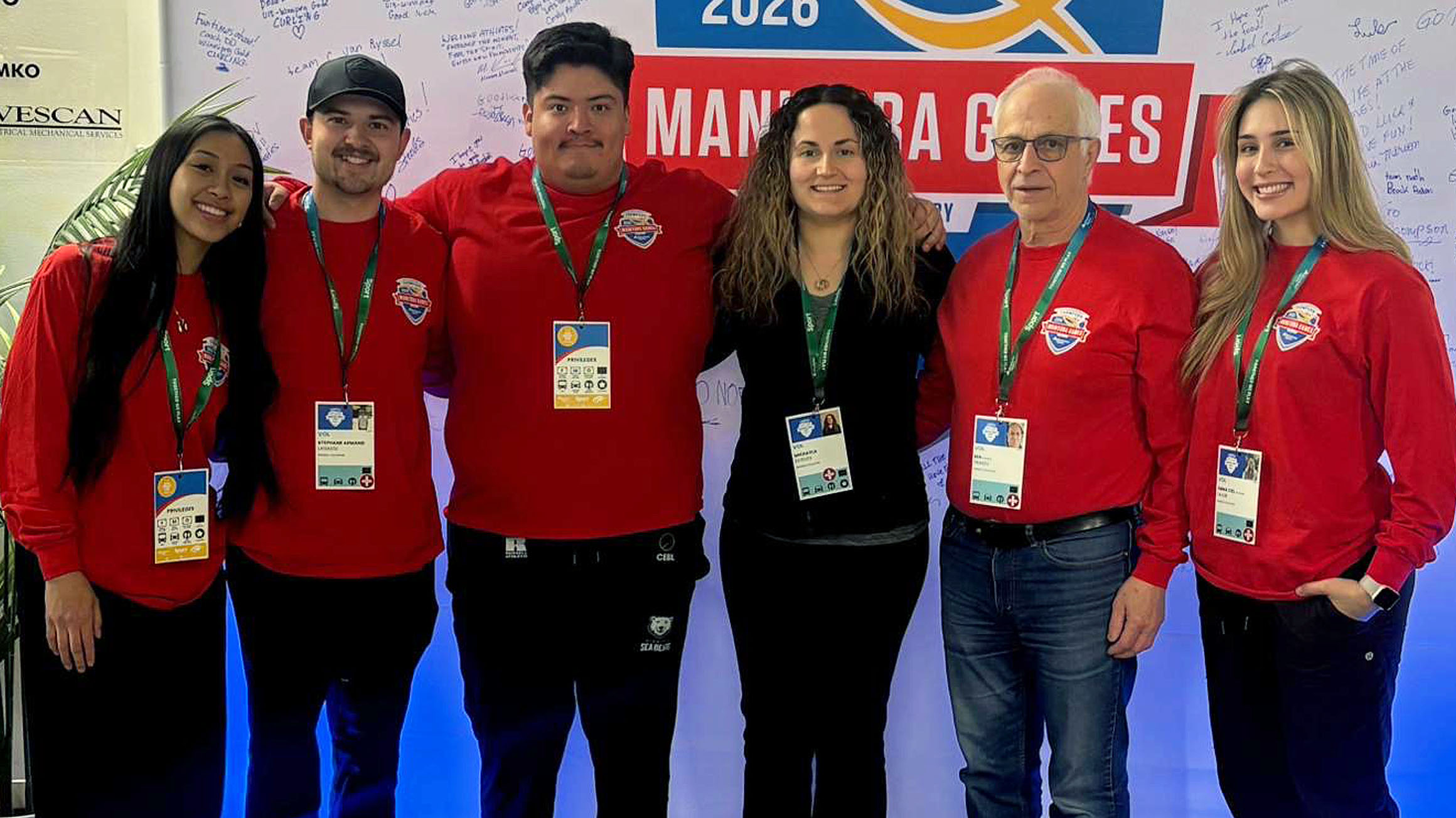 Six people, a mix of students and faculty members, stand in a row wearing red tops and lanyards at the Manitoba Games.