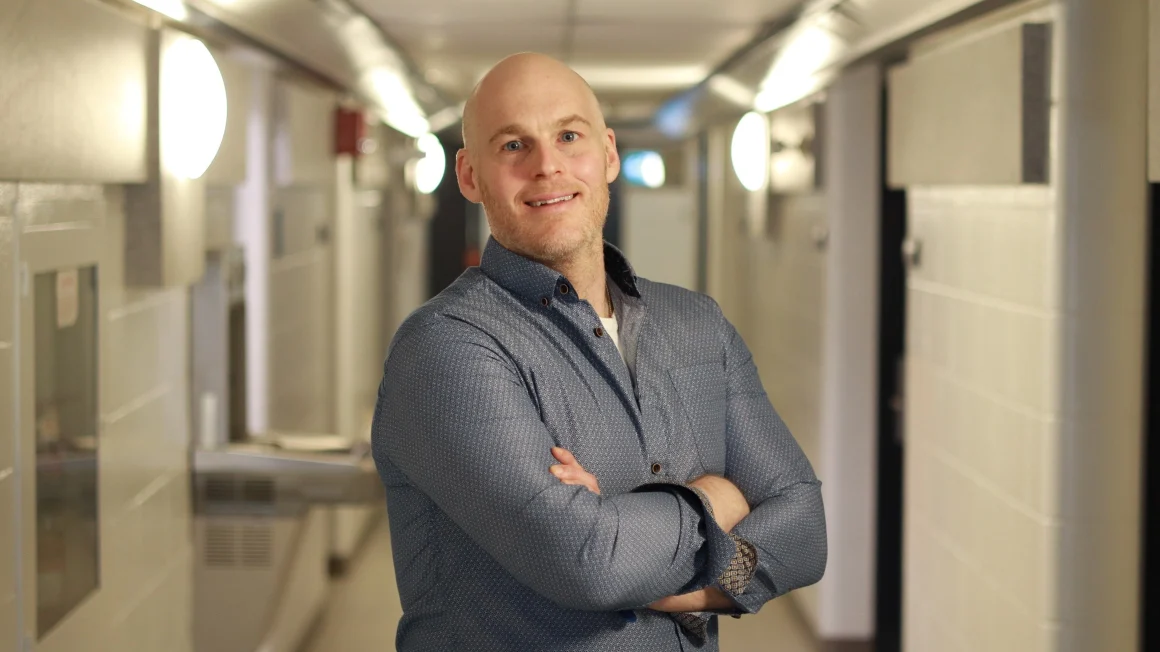 A smiling male professor wearing a dark blue patterned shirt stands in a university hallway with his arms folded.