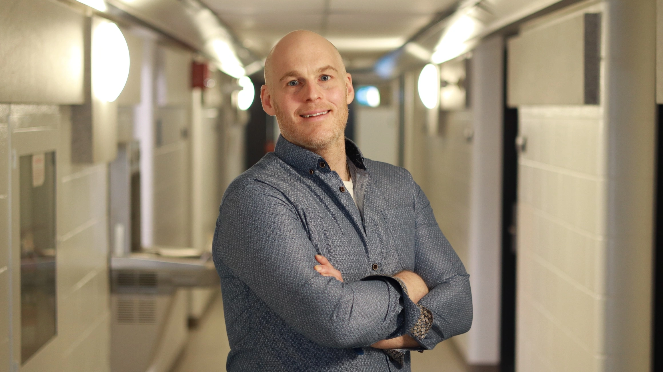 A smiling male professor wearing a dark blue patterned shirt stands in a university hallway with his arms folded.