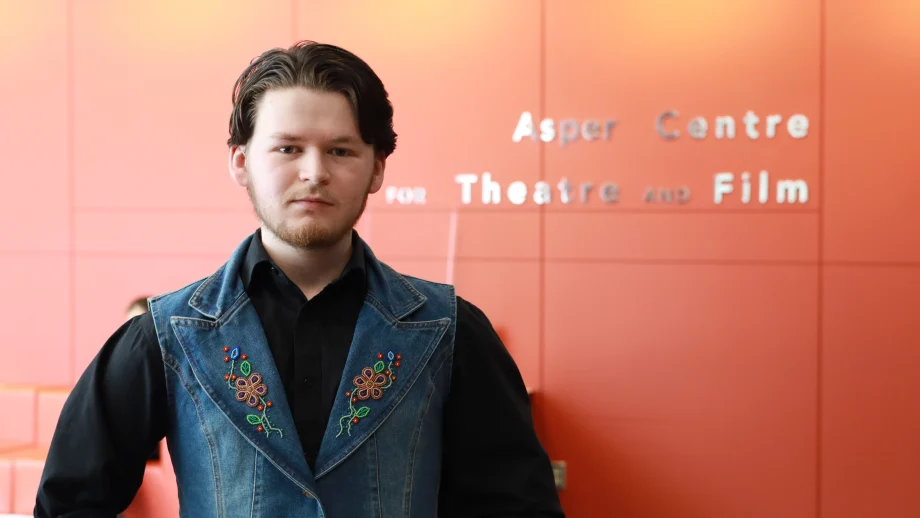 A male student with dark brown hair and an embroidered denim vest stands in front of a red wall with lettering that reads Asper Centre for Theatre and Film."