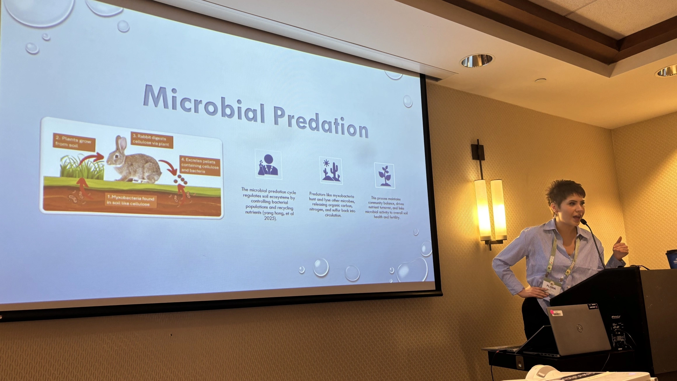 A female student with short brown hair in a white and blue striped shirt presents at a lectern beside a large drop-down screen displaying research slides.